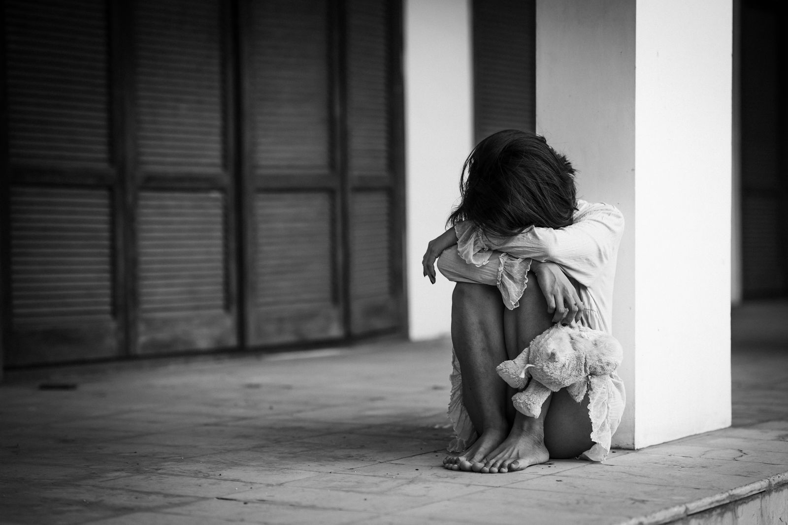 A young girl sitting alone on a porch, face hidden, holding a stuffed animal